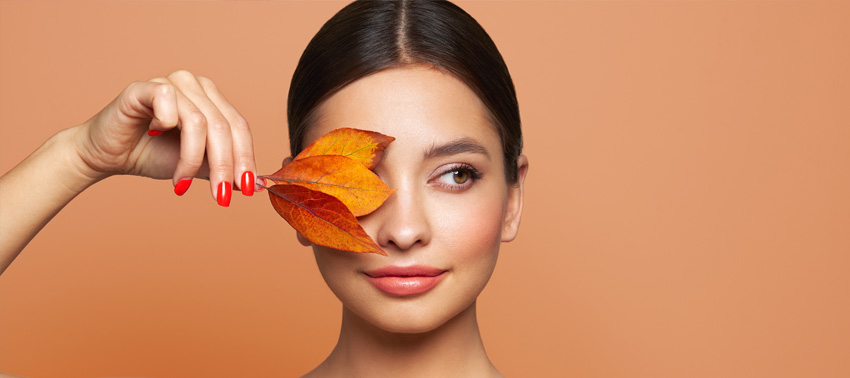 Woman with glowing skin holding autumn leaves over one eye against a warm orange background